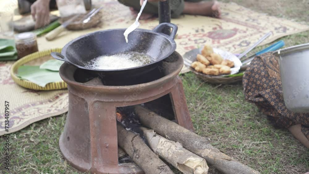 A woman dressed in traditional Javanese clothing is cooking Gorengan dough using a firewood stove and a frying pan. Javanese fries. A woman dressed in traditional Javanese clothing is cooking Gorengan dough using a firewood stove and a frying pan. Javanese fries.