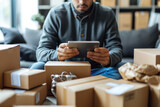 A focused man sits amidst an array of cardboard boxes, managing logistics on his tablet in a home business setting.