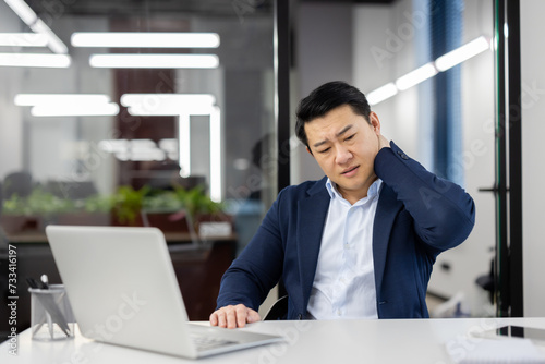 Overworked overtired asian man in business suit at workplace inside office, businessman has severe pain in neck, massaging muscle , long time sitting work with laptop, daily business of financier. photo
