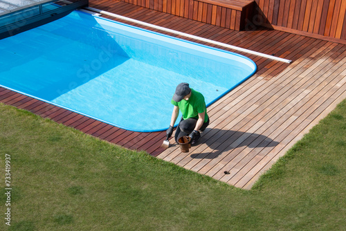 Exterior wooden pool deck painting, above view, person applying refreshing coat to poolside wood decking