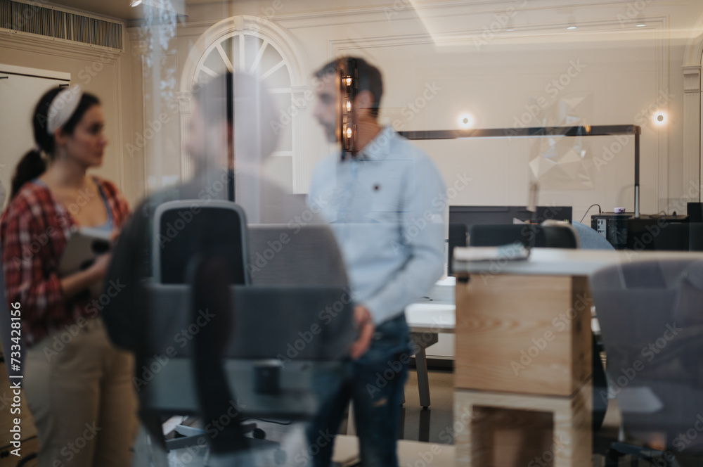 Two young professionals engaging in a discussion in a well-lit contemporary workspace with blurred glass foreground adding depth. Two young professionals engaging in a discussion in a well-lit contemporary workspace with blurred glass foreground adding depth.