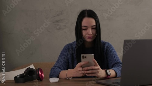 A young girl with a smartphone sits at a table with a laptop