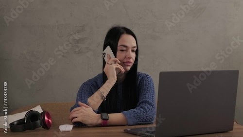 The girl is talking on the phone while sitting at the laptop in the room