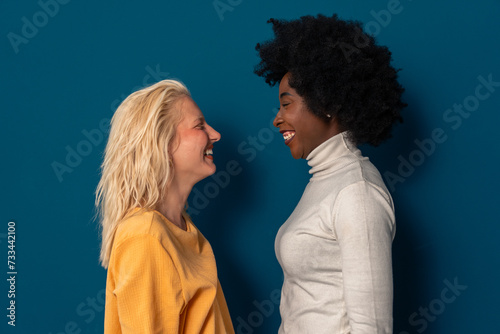 Two cheerful young women with different skin tones smiling at each other while standing together in a studio against a blue background.