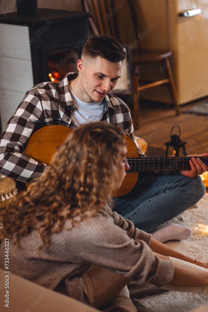 Obraz premium Young handsome man musician playing guitar instrumental music for his happy beautiful girlfriend near fireplace in a countryside cottage house, mountain chalet. Winter or autumn, cozy home atmosphere