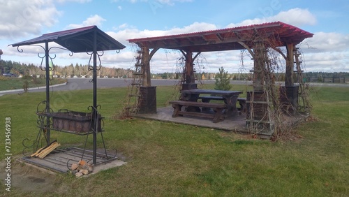 A metal brazier with a canopy and a wooden gazebo with benches, tables and a plastic roof stand on a planked area surrounded by lawns. There is a paved road, parking and mixed woods nearby