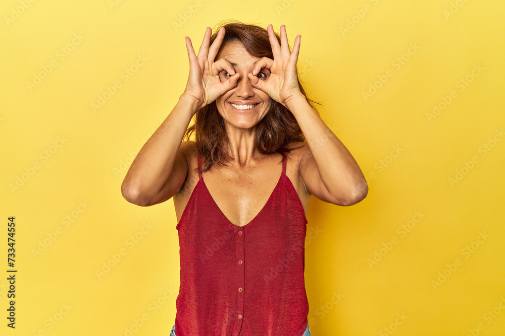 Middle-aged woman on a yellow backdrop showing okay sign over eyes Middle-aged woman on a yellow backdrop showing okay sign over eyes