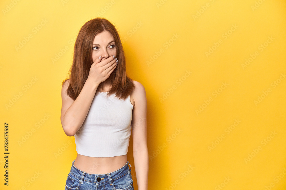 Redhead in white tank top, relaxed studio pose thoughtful looking to a copy space covering mouth with hand. Redhead in white tank top, relaxed studio pose thoughtful looking to a copy space covering mouth with hand.