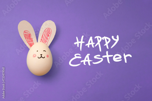 Chicken egg with cute rabbit face and bunny ears on lavender background. Food photo for Easter holiday. Greeting card. Happy Easter handwritten white text. Preparation for celebration. Family holiday