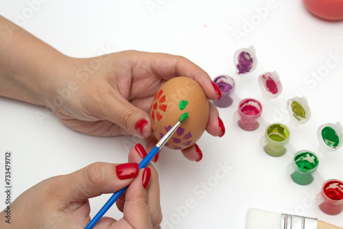 Woman draws flower and leaves on beige boiled chicken egg. Female hand holds brush. Preparation for holiday of Happy Easter. Paints and paper on white background. Artist with red manicure. Springtime 