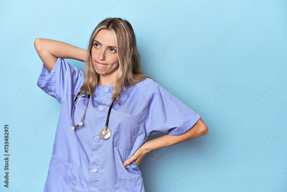 Young caucasian nurse in blue studio touching back of head, thinking and making a choice. Young caucasian nurse in blue studio touching back of head, thinking and making a choice.