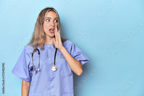 Young caucasian nurse in blue studio is saying a secret hot braking news and looking aside photo