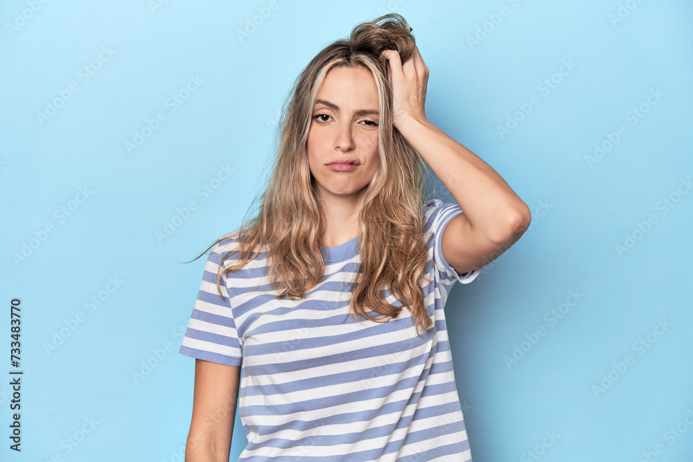 Blonde young caucasian woman in blue studio tired and very sleepy keeping hand on head.