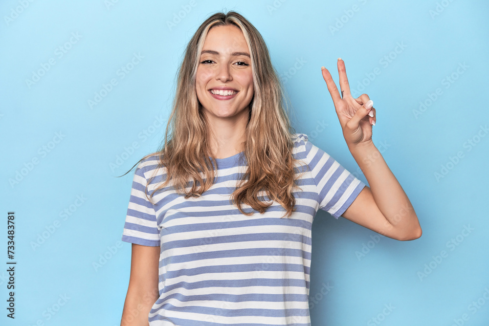 Blonde young caucasian woman in blue studio joyful and carefree showing a peace symbol with fingers. Blonde young caucasian woman in blue studio joyful and carefree showing a peace symbol with fingers.