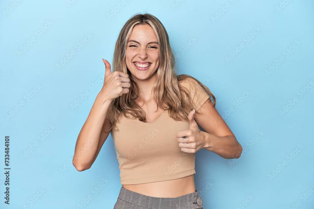 Fototapeta premium Blonde young caucasian woman in blue studio raising both thumbs up, smiling and confident.