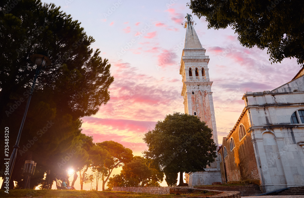 Rovinj, Istria, Croatia. Sunset in medieval old village town of Rovigno, tower Church Saint Euphemia during evening sunset. Picturesque scenic sky with clouds sun rays lighting through the trees.