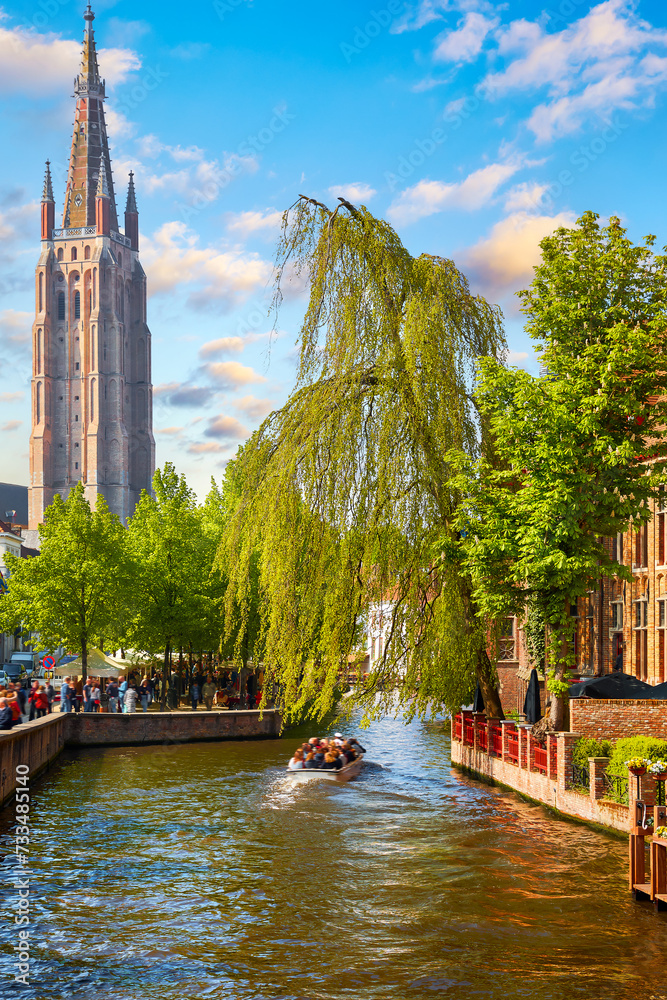 Fototapeta premium Bruges, Belgium. View at the Bell tower of church Our Lady in bruges old town. Channel Brugge with touristic boat and green trees during sunny summer day.