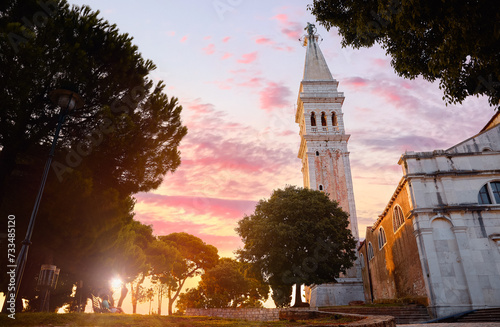 Rovinj  Istria  Croatia. Sunset in medieval old village town of Rovigno  tower Church Saint Euphemia during evening sunset. Picturesque scenic sky with clouds sun rays lighting through the trees.