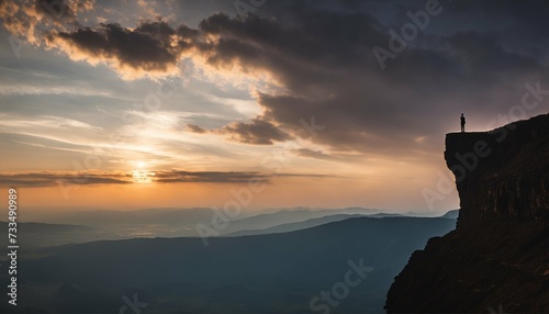 Silhouette of a person standing atop a cliff observing the sun rising amidst the clouds