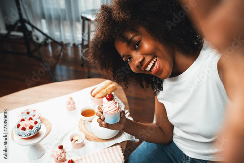 Beautiful young African blogger presenting piece of cupcake in concept special cuisine with selfie on smartphone. Content creating of social media with favorite sweets bakery dish. Tastemaker. photo