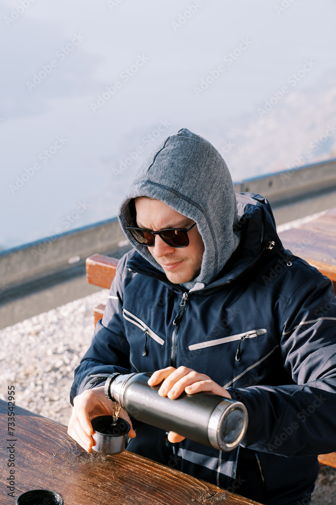 Man in a jacket with a pulled hood pours coffee from a thermos into a mug while sitting at a table in the mountains