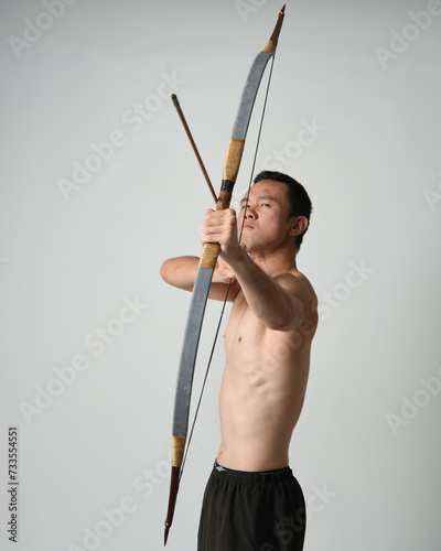 Full length portrait of fit  asian male model,  Holding hunting bow and arrow archery weapon, standing in warrior training action pose, isolated on white studio background.