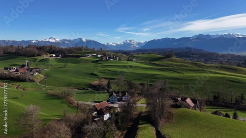 Beautiful green hills with small swiss village during sunny day. Panorama view on Glarner Alps in background. Snowy peaks in summer. Aerial panorama forward flight.
