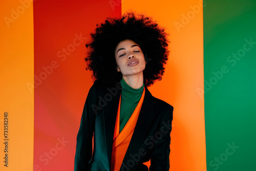 Stylish woman with afro, and office attire leaning on backdrop of warm orange and cool green