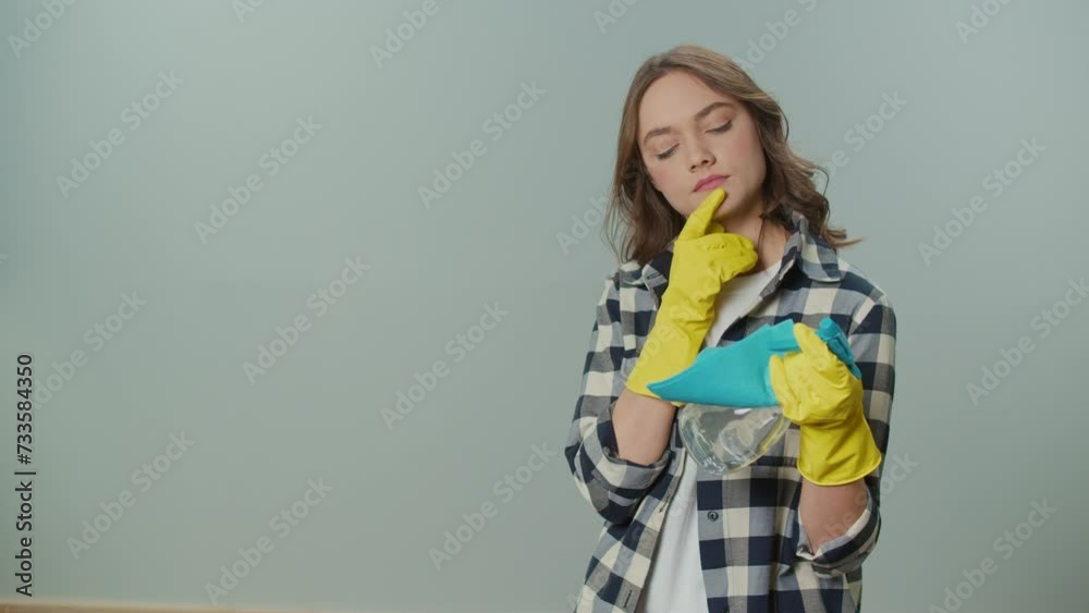 A Portrait of a Thoughtful Young Woman in Yellow Gloves,Holding a Cleaning Spray Bottle and Rag on the Gray Background.A Female Housewife Thinks Where to Start Cleaning. Hygiene and Safety Protocols. A Portrait of a Thoughtful Young Woman in Yellow Gloves,Holding a Cleaning Spray Bottle and Rag on the Gray Background.A Female Housewife Thinks Where to Start Cleaning. Hygiene and Safety Protocols.