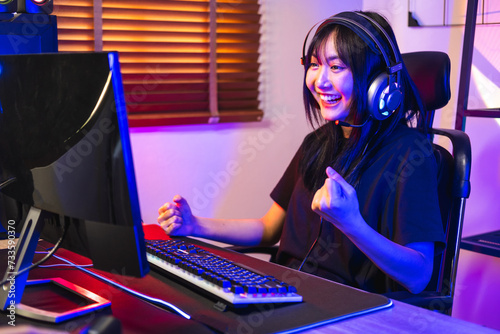 Happy asia girl gamer wear headphone competition play video game online with computer colorful neon light in living room at night