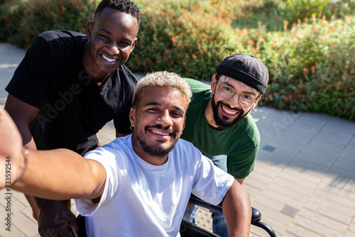 Young black man in a wheelchair using phone taking selfie together with male friends outdoors.