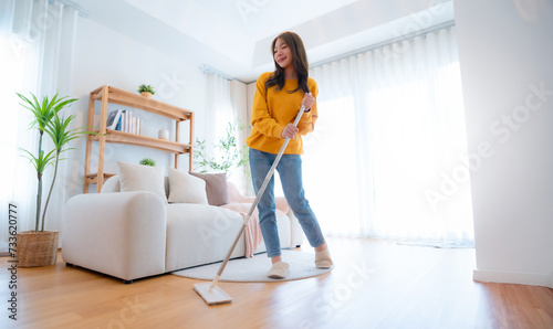 House cleaning with fun. Young adorable asian woman cleaning up living room, using mop as microphone, enjoying domestic work