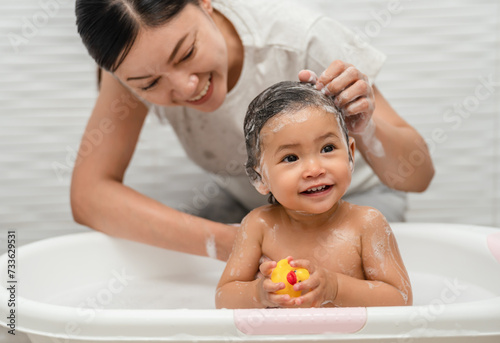mother bathing and washing her infant baby hair with shampoo while playing a rubber duck toy in bathtub photo