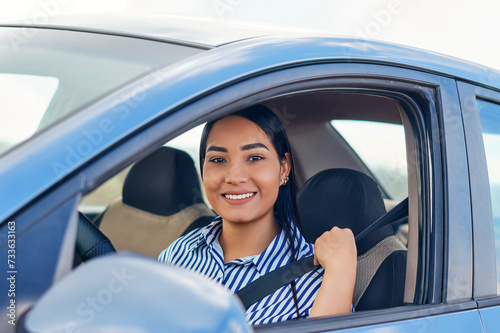 Smiling young latin woman looking at camera while putting on the seat belt before driving