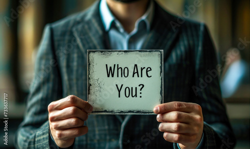 Close up of a man in a suit holding a card with Who Are You? printed on it, questioning identity and self perception