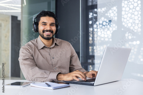 Carefree middle eastern guy in wireless headset typing on computer keyboard in modern office interior. Successful worker practising multitasking while listening to audiobook and answering emails. photo
