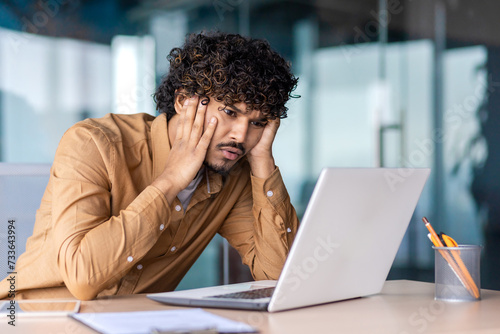 Confused muslim employee resting head on hands while looking at pc screen with incomprehension indoors. Curly male trainee desperately trying understanding project details on first working day. photo