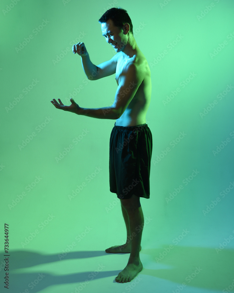 Close up portrait of fit asian male model, shirtless with muscles.  gestural ti chi inspired posing with arms reaching out  Isolated on a moody dark green studio background with silhouette.
