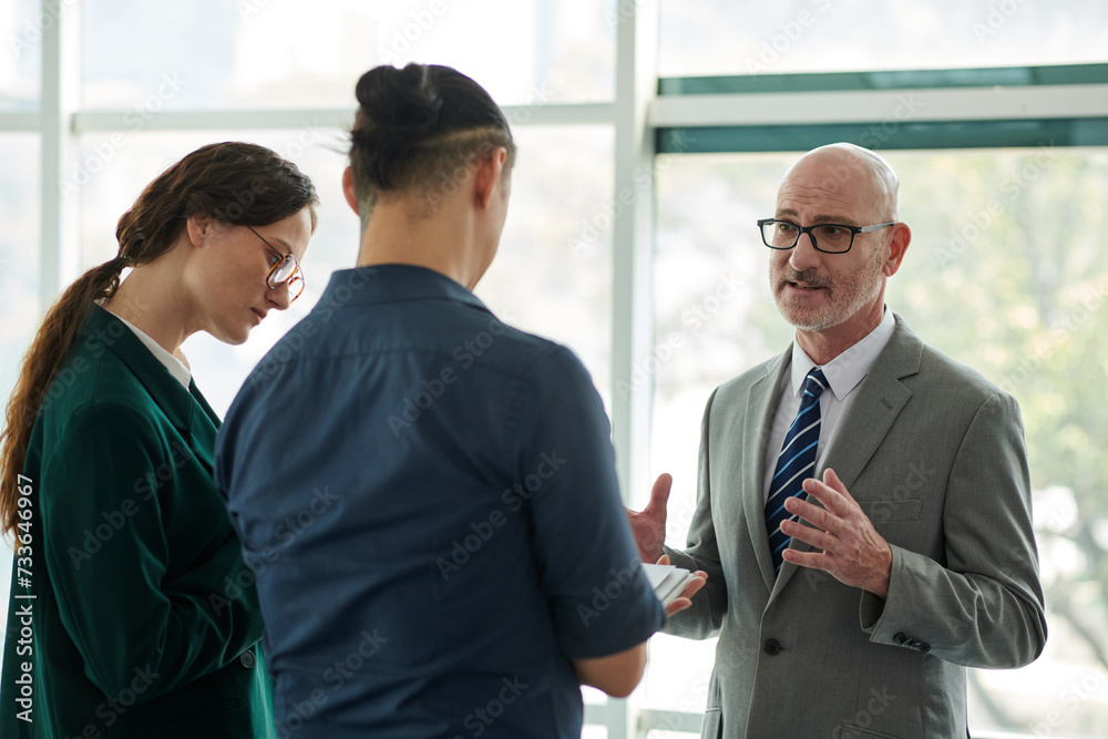 Confident director of business company in formalwear explaining working points to young subordinates at meeting in office center Confident director of business company in formalwear explaining working points to young subordinates at meeting in office center
