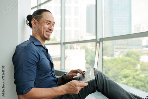 Happy young Asian male solopreneur with laptop typing on keyboard and looking at screen while sitting in front of large window photo
