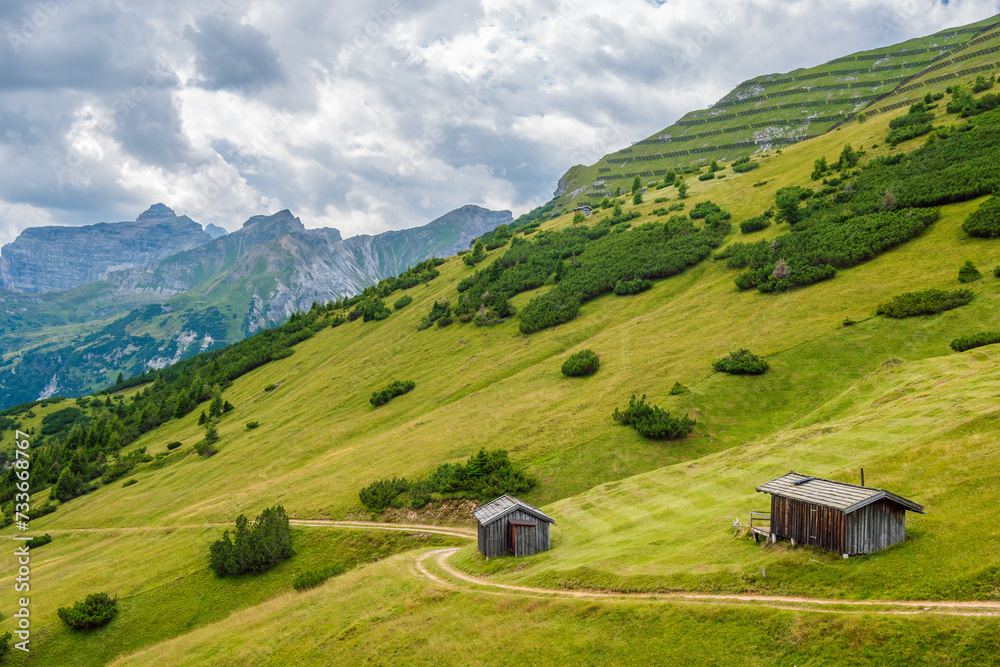 Picturesque summer mountain landscape from Stubai Alps, Tyrol, Austria. A green mountainside, two small wooden houses and a road winding past them, scenic alpine peak and cloudy sky in background.