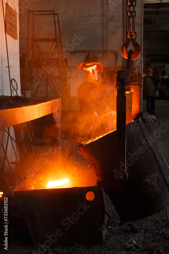 Fototapeta Naklejka Na Ścianę i Meble -  Worker in protective gear pours molten metal at steel mill. Industrial foundry scene showcasing manufacturing process. High-temperature metallurgical procedure by metalworker in safety equipment.