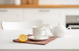 A cup of tea with lemon and a teapot on a white table. Kitchen with wooden empty countertop and brick wall in the background.