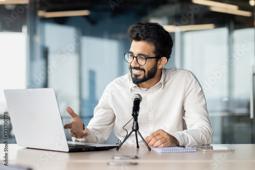 A smiling Indian young man is sitting in the office at a desk and talking through a microphone on a video call on a laptop. Conducts business training, holds a conference, online meeting