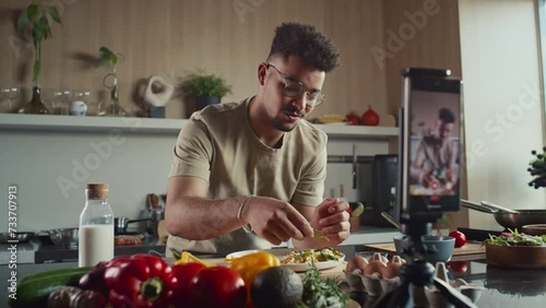 Young food blogger garnishing dish served on plate with fresh herbs and telling culinary hacks on camera of phone while filming cooking show in kitchen photo