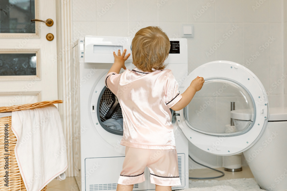 Rear view child girl little helper in pink pajama in laundry room near washing machine and dirty clothes in washing basket. washing and cleaning concept. daily routine. Copy space Rear view child girl little helper in pink pajama in laundry room near washing machine and dirty clothes in washing basket. washing and cleaning concept. daily routine. Copy space