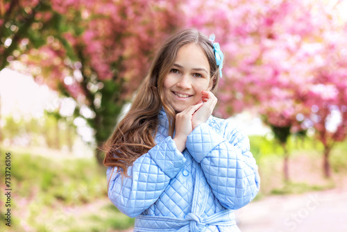 Young smile girl in a trench coat smiles standing at blossoming sakura garden. Outdoor fashion happy portrait little girl wearing stylish blue trench coat in blossom spring park. 