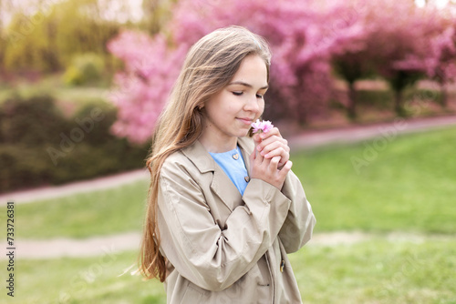 Outdoor fashion portrait little girl holding sakura flowers in his hands in blossom spring park. Young smile girl in a beige trench coat smiles and dreams standing at blossoming sakura garden. 