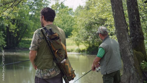Rear view of two fishermen on the river shore, using fishing rods with reels to catch a fish, medium shot. Concepts of enjoyment and recreation in nature.