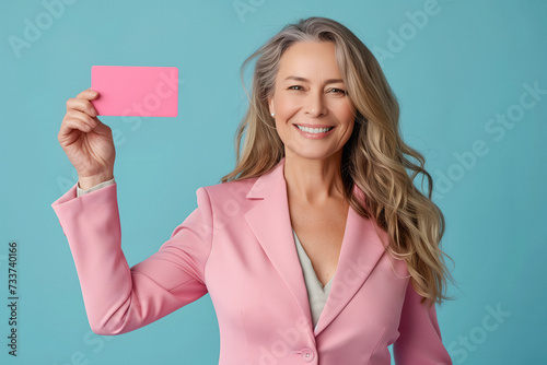 Positive middle-aged smiling woman in pink jacket is holding pink business or gift card, happy patient of mammalogical or gynecological clinic, plain blue background isolated photo